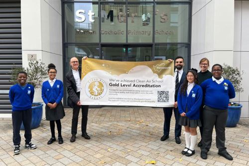 A group of seven people, including four pupils and three adults, stand outside St Mary's Roman Catholic Primary School holding a large banner. The banner reads: 'We've achieved Clean Air School! Gold Level Accreditation' and features the Wandsworth Council logo. The school's name is visible on the glass façade behind them.