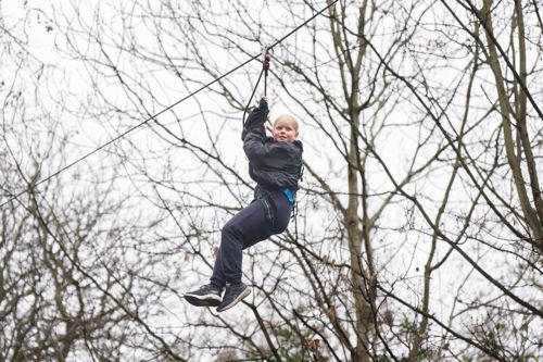A child wearing a dark jacket, blue harness, and dark athletic shoes is riding a zip line through a wooded area with bare trees. The individual is holding onto the zip line handle while suspended in mid-air.