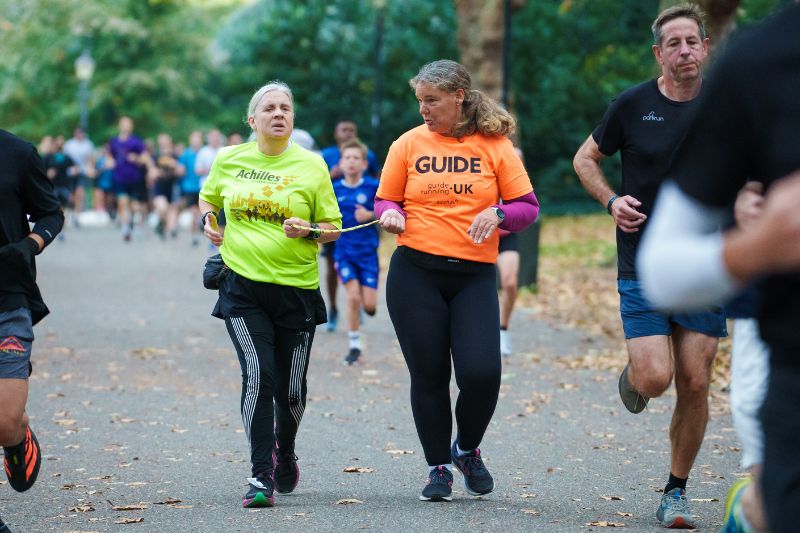 Two runners in the foreground participate in parkrun, with one wearing a yellow t-shirt and the other in an orange 'GUIDE' t-shirt. They are surrounded by other runners in a tree-lined park with autumn leaves on the ground.